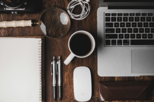 Overhead view of a desktop with a blank notebook, pens, coffee mug, laptop, and mouse—someone is ready to write!!